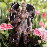 A highly detailed fantasy figurine depicted in intricate armor with dark wings stands amid blooming pink and white flowers. The figure has an intense expression and is surrounded by green foliage, creating a striking contrast between the natural setting and the otherworldly character.
