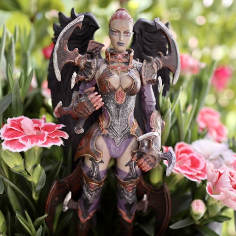 A highly detailed fantasy figurine depicted in intricate armor with dark wings stands amid blooming pink and white flowers. The figure has an intense expression and is surrounded by green foliage, creating a striking contrast between the natural setting and the otherworldly character.
