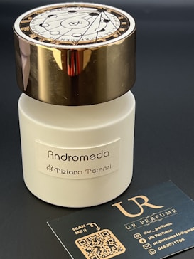 A white bottle of perfume labeled Andromeda by Tiziana Terenzi with a gold-colored cap. The bottle is accompanied by a black business card for UR Perfume, featuring QR codes and social media handles.