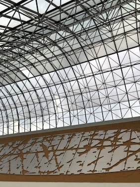 A geometric glass ceiling with a grid of black metal beams forms intricate patterns, allowing natural light to filter through. Below the glass ceiling, a decorative wall features abstract patterns in white with a brown background, adding an artistic touch to the architectural design.