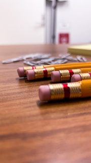 Four pencils with pink erasers are lying on a wooden table, closely aligned. In the background, there are blurred paper clips and a stack of sticky notes.