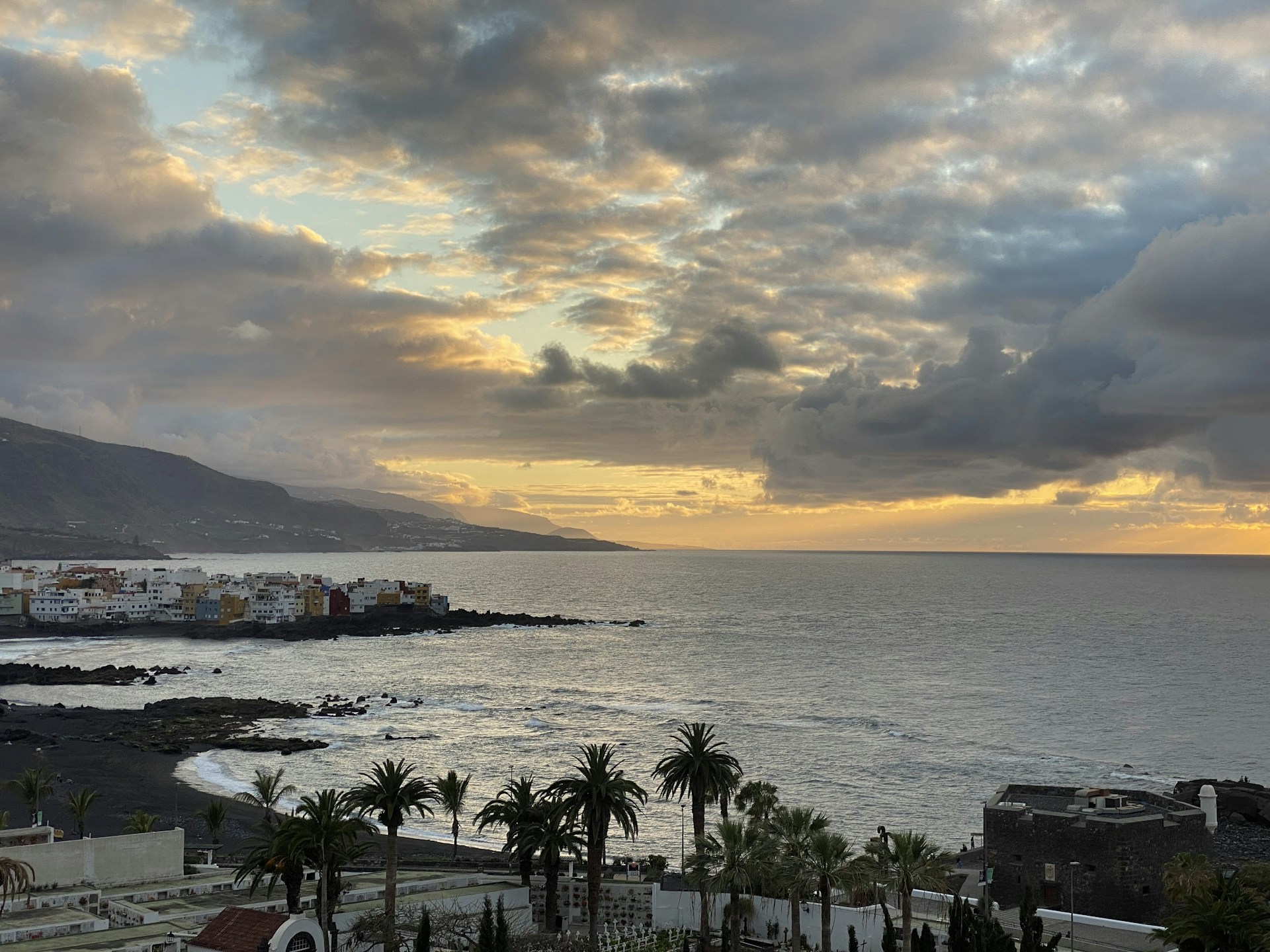 A coastal scene featuring a small town with colorful buildings situated near the ocean. The water has gentle waves, and the shoreline is lined with rocks. Tall palm trees are visible in the foreground, while a mountainous landscape can be seen in the distance. The sky is filled with dramatic clouds and a warm, golden sunset.