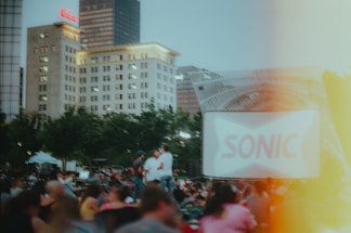 A large outdoor gathering during dusk with a crowd seated in front of a screen showing a blurred advertisement for Sonic. There are several tall buildings in the background, and the area is surrounded by trees. The mood is lively with people enjoying an open-air event.