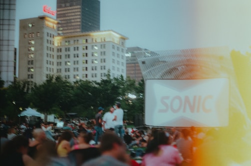 A large outdoor gathering during dusk with a crowd seated in front of a screen showing a blurred advertisement for Sonic. There are several tall buildings in the background, and the area is surrounded by trees. The mood is lively with people enjoying an open-air event.