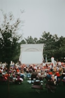 An outdoor setting with a large group of people seated in chairs facing a blank screen, suggesting a community event or outdoor movie night. Trees are visible in the background, and the overall scene is slightly blurred, conveying a relaxed atmosphere.