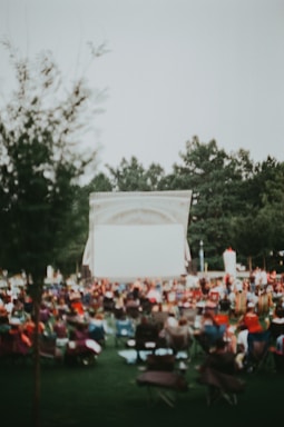 An outdoor setting with a large group of people seated in chairs facing a blank screen, suggesting a community event or outdoor movie night. Trees are visible in the background, and the overall scene is slightly blurred, conveying a relaxed atmosphere.