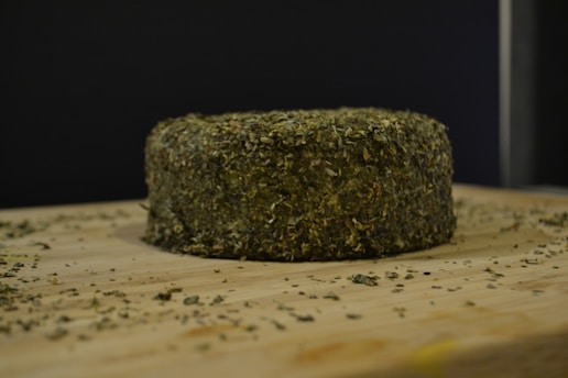 A round wheel of cheese coated with a dense layer of herbs is placed on a wooden surface, surrounded by scattered herbs. The background is dark, creating a contrast that highlights the cheese.