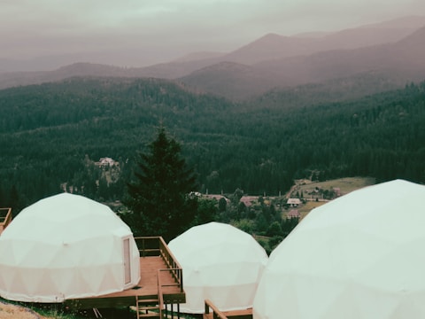 Geodesic domes are situated on a wooden deck overlooking a vast forested landscape with mountains in the background. The overcast sky and mist create a serene and tranquil ambiance.