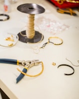 A workspace featuring various jewelry-making tools and materials. A spool of beige thread is placed upright next to a pair of pliers with blue and yellow handles. Several jewelry pieces, including bracelets and small metal components, are scattered on the white surface.