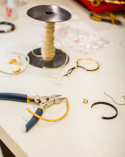 A workspace featuring various jewelry-making tools and materials. A spool of beige thread is placed upright next to a pair of pliers with blue and yellow handles. Several jewelry pieces, including bracelets and small metal components, are scattered on the white surface.