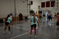 A group of people are playing volleyball indoors. The players are divided into two teams, actively engaging with the game as the ball is in the air. The gymnasium has wooden benches against the wall and a volleyball net dividing the courts. Some players are wearing casual sportswear, including t-shirts and shorts, and one player has bright pink knee-high socks.