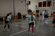 A group of people are playing volleyball indoors. The players are divided into two teams, actively engaging with the game as the ball is in the air. The gymnasium has wooden benches against the wall and a volleyball net dividing the courts. Some players are wearing casual sportswear, including t-shirts and shorts, and one player has bright pink knee-high socks.
