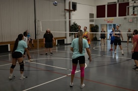 A group of people are playing volleyball indoors. The players are divided into two teams, actively engaging with the game as the ball is in the air. The gymnasium has wooden benches against the wall and a volleyball net dividing the courts. Some players are wearing casual sportswear, including t-shirts and shorts, and one player has bright pink knee-high socks.