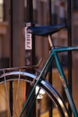 A close-up of a bicycle secured to a metal railing. A sticker on the railing reads 'J'EXISTE.' The bicycle is teal with curved handlebars and a steel frame. The background is softly blurred, creating a warm, evening city ambiance.