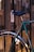 A close-up of a bicycle secured to a metal railing. A sticker on the railing reads 'J'EXISTE.' The bicycle is teal with curved handlebars and a steel frame. The background is softly blurred, creating a warm, evening city ambiance.