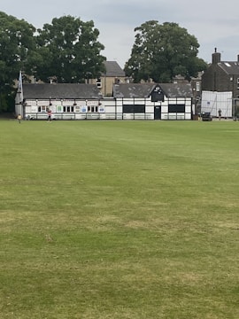 A large open field of green grass with a traditional black and white half-timbered building in the background. The structure seems to be a cricket pavilion, accompanied by a few people and trees surrounding the area.