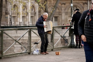 A street musician plays the accordion on a wooden bridge, with a basket on the ground for donations. Another person stands nearby, facing away and holding a bouquet of flowers. The background features classic architecture with arched windows, and a few pedestrians are visible.