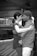 A black and white photograph of a man and a woman sharing an intimate embrace, standing close together in a dimly lit indoor setting. The woman, with dark hair pulled back, wears a patterned top, while the man sports a patterned shirt and a backwards cap. Their expressions suggest a deep emotional connection.