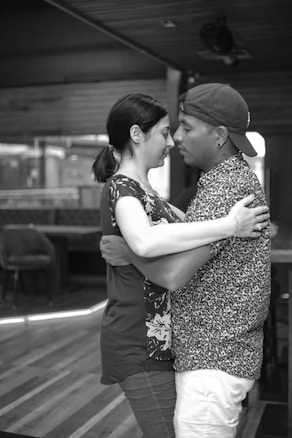 A black and white photograph of a man and a woman sharing an intimate embrace, standing close together in a dimly lit indoor setting. The woman, with dark hair pulled back, wears a patterned top, while the man sports a patterned shirt and a backwards cap. Their expressions suggest a deep emotional connection.