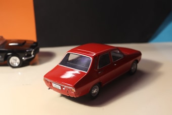 A model of a red vintage car is prominently displayed on a light surface with an orange and black background. To the left, a black model car is partially visible, adding contrast.