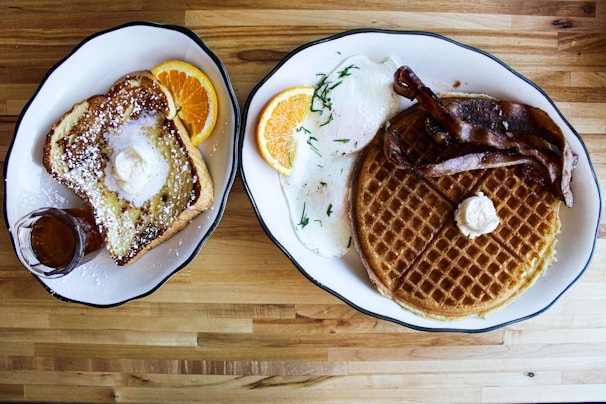 Two plates on a wooden table. The left plate features French toast topped with butter and powdered sugar, accompanied by orange slices and a glass of syrup. The right plate contains a waffle with butter, crispy bacon strips, over-easy eggs garnished with herbs, and an orange slice.