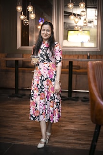 A woman in a floral dress holds a cocktail glass while standing in a stylishly decorated room with warm lighting and modern hanging light fixtures. The background features a TV screen and cushioned bar seating.