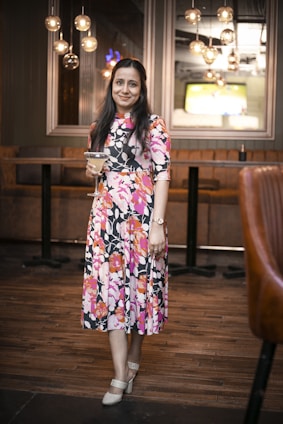 A woman in a floral dress holds a cocktail glass while standing in a stylishly decorated room with warm lighting and modern hanging light fixtures. The background features a TV screen and cushioned bar seating.