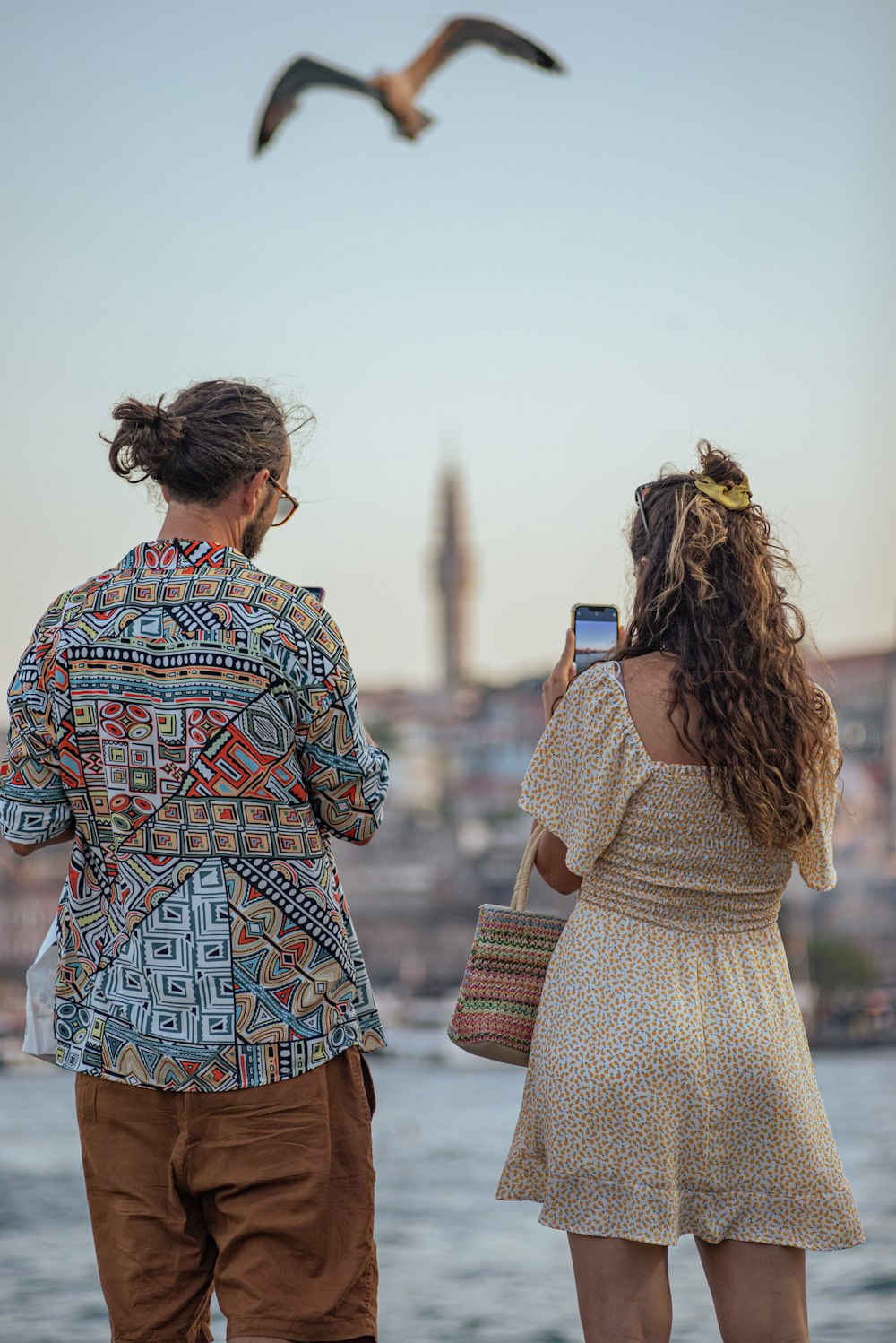 a couple of people stand on a beach looking at a bird flying