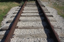Old, weathered train tracks run through a rocky terrain surrounded by patches of grass. The wooden planks are worn and the metal tracks are rusted, indicating age and disuse. Sunlight casts shadows across the railroad, enhancing the texture of the gravel and wood.
