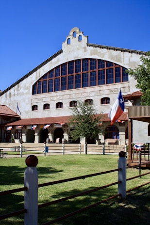 A historic building with an arched facade featuring large windows and a Texas flag flying in front. The building is decorated with bunting and surrounded by a wooden fence. The lawn is well-maintained and the sky is clear and blue.