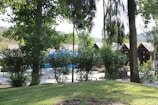 A tranquil outdoor setting with lush greenery and trees surrounding a small cluster of wooden cabins with pointed roofs. A vintage-style lamp post stands amidst the foliage, and a blue canopy is visible in the background, while a well-kept lawn covers the foreground.