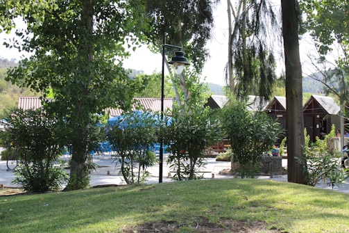 A tranquil outdoor setting with lush greenery and trees surrounding a small cluster of wooden cabins with pointed roofs. A vintage-style lamp post stands amidst the foliage, and a blue canopy is visible in the background, while a well-kept lawn covers the foreground.