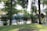 A tranquil outdoor setting with lush greenery and trees surrounding a small cluster of wooden cabins with pointed roofs. A vintage-style lamp post stands amidst the foliage, and a blue canopy is visible in the background, while a well-kept lawn covers the foreground.