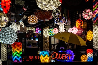 A vibrant display of colorful geometric pendant lamps hanging in a shop with varied designs and shapes. The scene is filled with multiple bright, intricate light fixtures, some spherical and interwoven. A neon sign with the word 'Ouvert' is illuminated, while a yellow umbrella with writing on it is visible below. The setting is an indoor market stall or shop with reflective, dark surfaces.