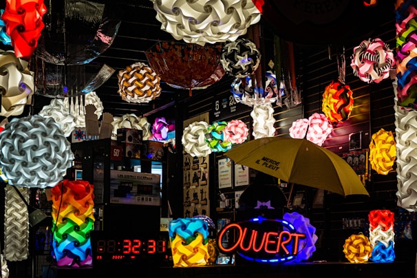 A vibrant display of colorful geometric pendant lamps hanging in a shop with varied designs and shapes. The scene is filled with multiple bright, intricate light fixtures, some spherical and interwoven. A neon sign with the word 'Ouvert' is illuminated, while a yellow umbrella with writing on it is visible below. The setting is an indoor market stall or shop with reflective, dark surfaces.