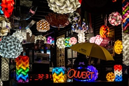 A vibrant display of colorful geometric pendant lamps hanging in a shop with varied designs and shapes. The scene is filled with multiple bright, intricate light fixtures, some spherical and interwoven. A neon sign with the word 'Ouvert' is illuminated, while a yellow umbrella with writing on it is visible below. The setting is an indoor market stall or shop with reflective, dark surfaces.