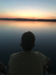 Back view of a person sitting peacefully by a calm lake at sunset, immersed in thought.