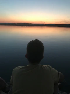 Back view of a person sitting peacefully by a calm lake at sunset, immersed in thought.