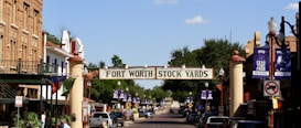 A street scene featuring a historic district with a prominent 'Fort Worth Stockyards' sign arching over the road. The street is lined with old-fashioned buildings, street lamps, and banners. Vehicles are parked along the street, and the atmosphere suggests a blend of history and tourism.