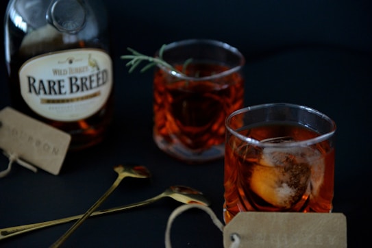 A bottle of Wild Turkey Rare Breed whiskey is placed on a dark surface alongside two glasses filled with amber-colored liquid, ice, and a sprig of rosemary. Next to them are two gold-colored spoons with tags attached.