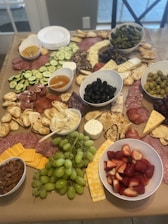 A large charcuterie board is adorned with an assortment of cheeses, various types of sliced meats, crackers, breads, and fruits. Bowls contain items such as olives, strawberries, and almonds, while a spread of cucumbers, green grapes, and sliced cheeses adds variety. Dips and spreads, including hummus, accompany the selection, which is meticulously arranged on a wooden table.