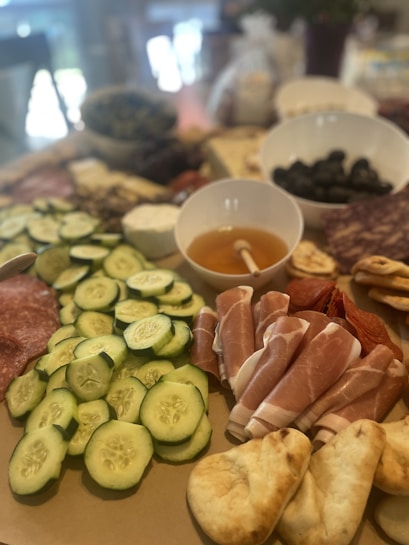 A variety of fresh and cured foods are arranged on a wooden board. Sliced cucumbers, cured meat, and prosciutto are prominently displayed alongside small rounds of bread. Bowls containing olives and what appears to be honey with a dipper are included in the spread.
