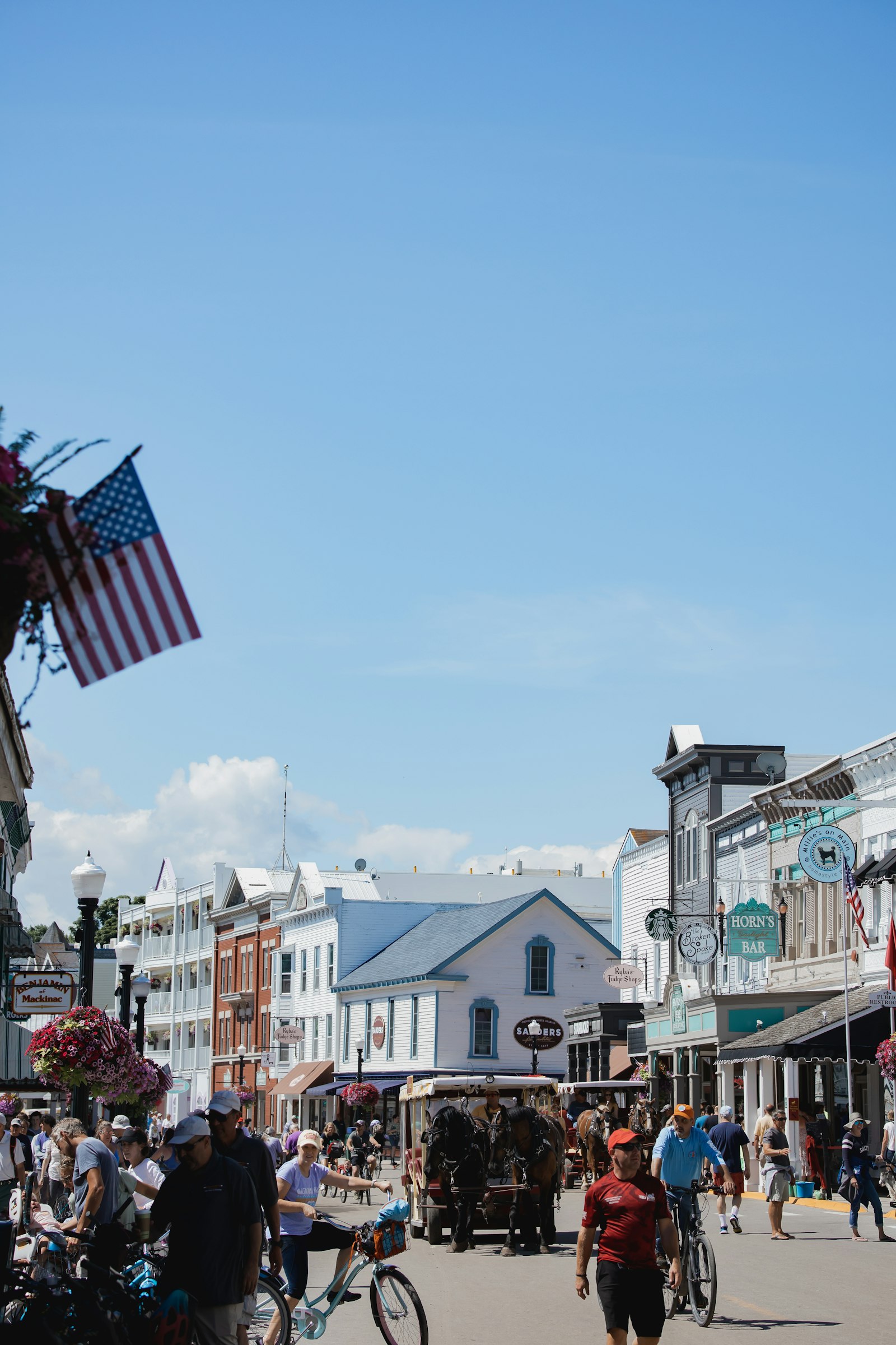 A small-town main street busy with pedestrians and cyclists, an American flag flying and a horse-drawn wagon in the distance