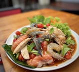 A vibrant plate of seafood and fresh vegetables presented on a white dish. The dish includes raw shrimp, salmon slices, crab, cherry tomatoes, green beans, and mint leaves, all lightly sprinkled with sesame seeds. The ingredients are arranged atop a green leafy base, with an appetizing sauce visible at the base.