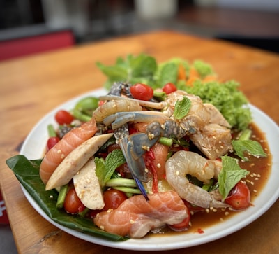 A vibrant plate of seafood and fresh vegetables presented on a white dish. The dish includes raw shrimp, salmon slices, crab, cherry tomatoes, green beans, and mint leaves, all lightly sprinkled with sesame seeds. The ingredients are arranged atop a green leafy base, with an appetizing sauce visible at the base.