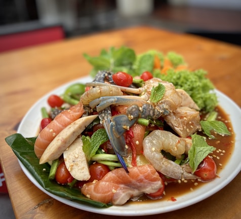 A vibrant plate of seafood and fresh vegetables presented on a white dish. The dish includes raw shrimp, salmon slices, crab, cherry tomatoes, green beans, and mint leaves, all lightly sprinkled with sesame seeds. The ingredients are arranged atop a green leafy base, with an appetizing sauce visible at the base.