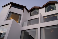 Contemporary architecture featuring a white multi-level building with large windows and dark slanted roofs. The structure is modern and minimalistic, exuding a sense of sophistication. Glass balconies and drapes can also be seen through the expansive windows.