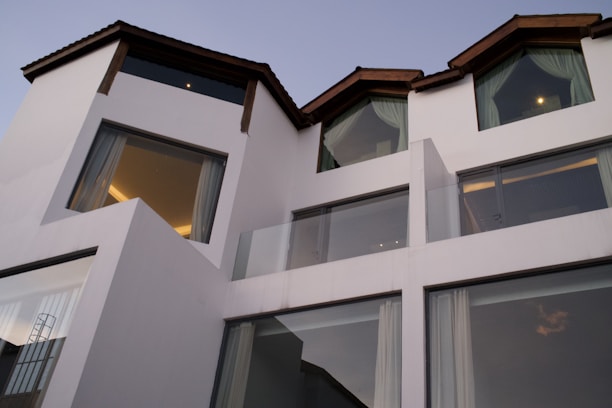 Contemporary architecture featuring a white multi-level building with large windows and dark slanted roofs. The structure is modern and minimalistic, exuding a sense of sophistication. Glass balconies and drapes can also be seen through the expansive windows.