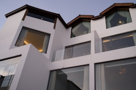 Contemporary architecture featuring a white multi-level building with large windows and dark slanted roofs. The structure is modern and minimalistic, exuding a sense of sophistication. Glass balconies and drapes can also be seen through the expansive windows.