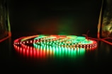 A series of LED light strips are coiled on a dark surface, illuminated in alternating red and green colors. The glowing lights create reflections on the glossy surface below, providing a striking contrast against the dark background.